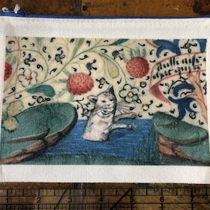 Może przedstawiać: Biała płócienna kosmetyczka z niebieskim zamkiem błyskawicznym. Kosmetyczka przedstawia vintage'ową ilustrację białego kota na tle kwiatów i napis "dullk nyfe" pisany kursywą.