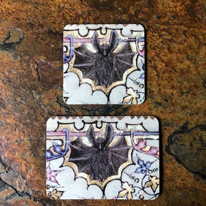 May include: Two square coasters with a white background and a black bat illustration. The bat is surrounded by gold and purple floral designs. The coasters have a vintage look.