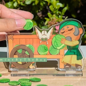 May include: A clear acrylic charm featuring a cartoon character with a green hat and outfit, hugging a cabbage. A wooden cart with cabbages and a wheel is behind the character. The charm has the text "Jasmine By The Bay".