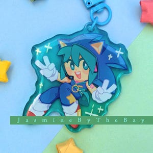 May include: A blue and green character with a star on their chest and a peace sign on each hand. The character is on a clear blue background with white sparkles. The character is a keychain with a blue and white string.