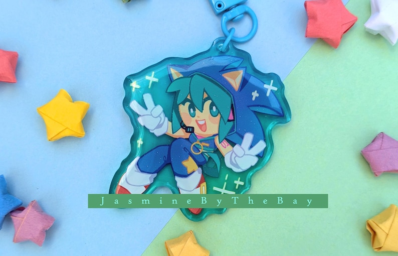 May include: A blue and green character with a star on their chest and a peace sign on each hand. The character is on a clear blue background with white sparkles. The character is a keychain with a blue and white string.