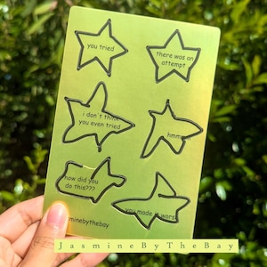 Könnte beinhalten: Ein metallisch-grünes Stickerbogen mit sechs sternförmigen Sprechblasen. Jede Blase enthält einen anderen Satz in schwarzer Schrift, darunter "you tried" und "there was an attempt". Der Markenname "Jasmine By The Bay" steht unten.