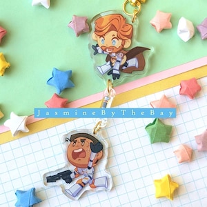 May include: Two cartoon character keychains with gold-colored hardware. One keychain features a character with orange hair and a brown cape. The other keychain features a character holding a weapon. Colorful origami stars surround the keychains. The text "Jasmine By The Bay" is visible.