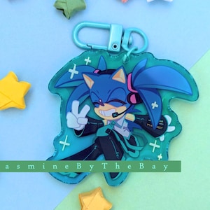 May include: A blue and green character keychain with a blue hedgehog wearing headphones and a green jacket. The keychain has a clear background with white plus signs. The text "Jasmine By The Bay" is visible at the bottom of the keychain.