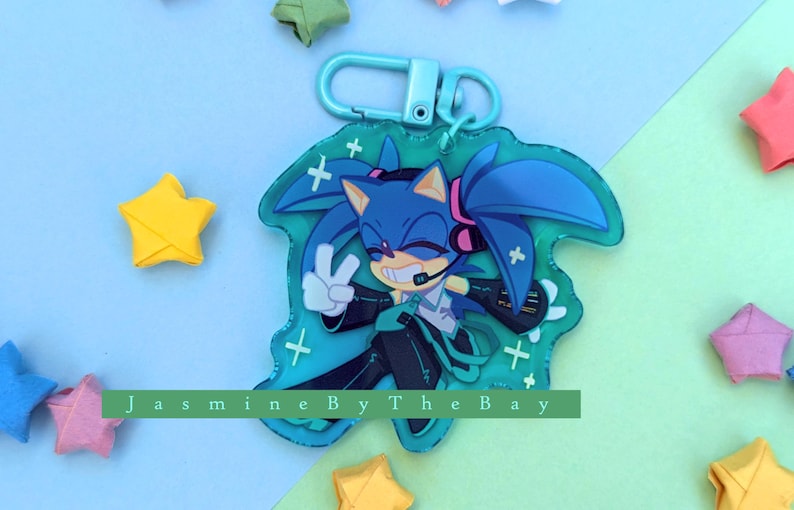 May include: A blue and green character keychain with a blue hedgehog wearing headphones and a green jacket. The keychain has a clear background with white plus signs. The text "Jasmine By The Bay" is visible at the bottom of the keychain.