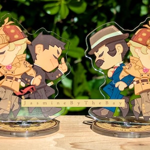 May include: Two acrylic character stands featuring two cartoon characters. The characters are dressed in detective outfits and are holding cups of coffee. The stands are on a wooden surface. The text "Jasmine By The Bay" is visible on the stands.