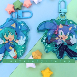 May include: Two blue and green character keychains with a sparkly background. The characters are from the Sonic the Hedgehog franchise. The keychains are attached to silver key rings.