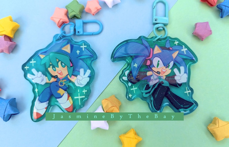 May include: Two blue and green character keychains with a sparkly background. The characters are from the Sonic the Hedgehog franchise. The keychains are attached to silver key rings.
