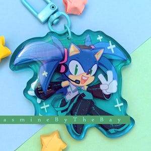 May include: A blue and green character with blue hair and a green outfit, wearing headphones, with a peace sign, on a clear acrylic keychain with a silver metal ring.