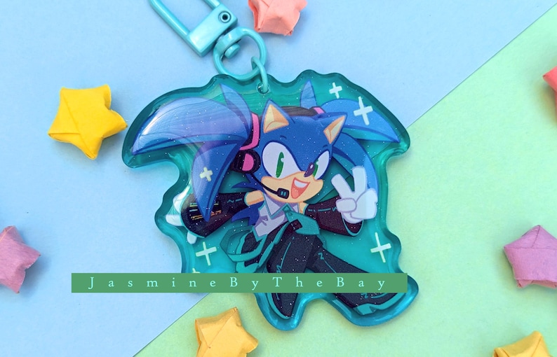 May include: A blue and green character with blue hair and a green outfit, wearing headphones, with a peace sign, on a clear acrylic keychain with a silver metal ring.