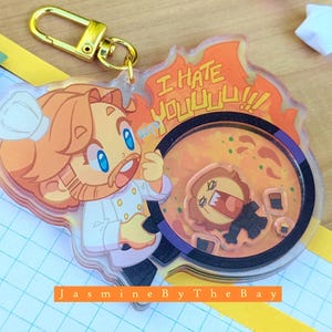 May include: A clear acrylic keyring featuring a cartoon chef with orange hair and a white chef's hat. The keyring has the text "I HATE YOU!!!" and a cartoon image of a chef in a pan. The keyring is attached to a gold-coloured clasp.