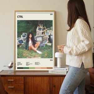 Könnte beinhalten: Ein gerahmtes Poster mit dem Albumcover von SZA's "CTRL". Das Poster hat einen beigen Hintergrund mit dem Albumtitel und dem Jahr. Das Bild zeigt SZA, die vor einem Haufen alter Computermonitore sitzt. Das Poster wird auf einem Holzschrank ausgestellt.