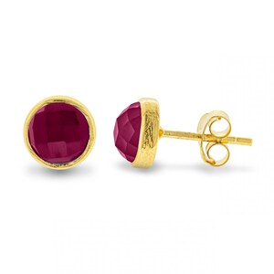 May include: A pair of gold stud earrings with a round, faceted, burgundy gemstone.