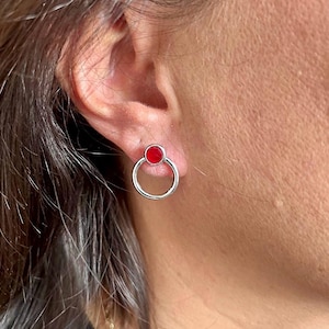 Könnte beinhalten: Ein silberner Ohrstecker mit einem roten, kreisförmigen Stein über einem silbernen Ring. Der Ohrring wird an einem Ohr gezeigt, wobei der Fokus auf dem Schmuck liegt. Das Design ist schlicht und modern.