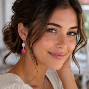 May include: A pair of gold-toned drop earrings featuring a small, round, white stone at the top and a larger, teardrop-shaped, vibrant pink textured stone at the bottom, suspended from a simple gold hook wire design.
