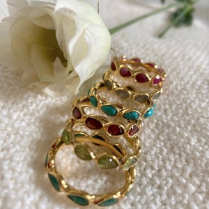 May include: A set of five gold rings with different coloured gemstones. The rings are stacked on top of each other, with the largest ring at the bottom and the smallest ring at the top. The gemstones are turquoise, green, red, and grey.