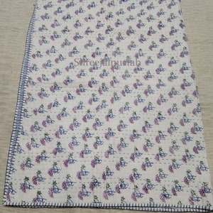 Pode incluir: Colcha de algod&atilde;o branco com um padr&atilde;o repetitivo de desenhos florais roxos e azuis. A colcha tem um fundo branco e &eacute; costurada &agrave; m&atilde;o com fio azul claro. A colcha &eacute; bordada com uma guarni&ccedil;&atilde;o azul escura.