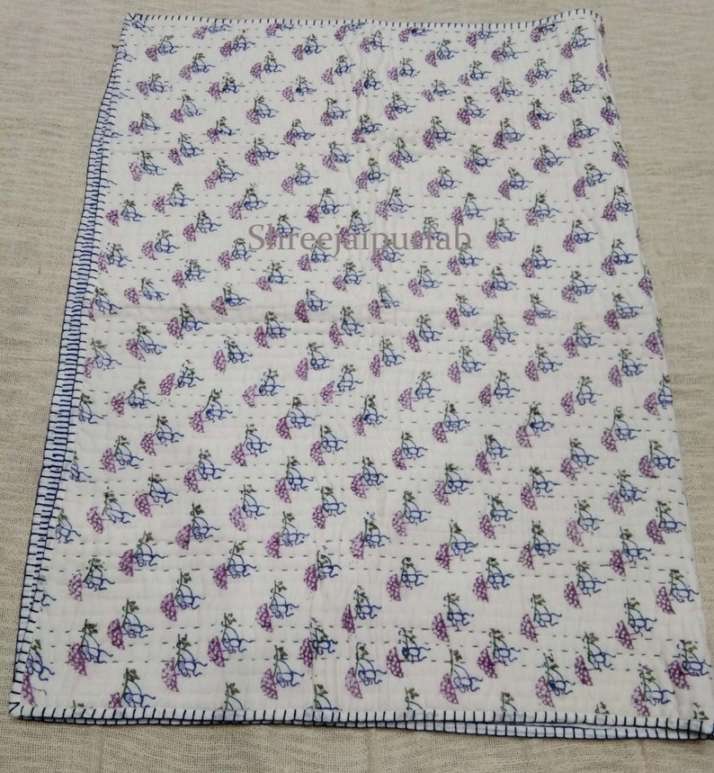 Pode incluir: Colcha de algod&atilde;o branco com um padr&atilde;o repetitivo de desenhos florais roxos e azuis. A colcha tem um fundo branco e &eacute; costurada &agrave; m&atilde;o com fio azul claro. A colcha &eacute; bordada com uma guarni&ccedil;&atilde;o azul escura.