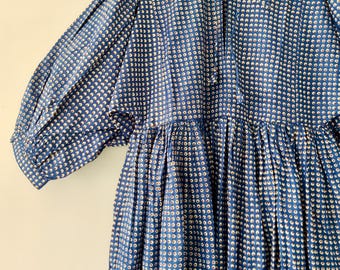 Puede incluir: Vestido azul con estampado de lunares blancos. El vestido tiene cuello redondo, mangas largas y cintura fruncida. Cuelga de una percha de madera.