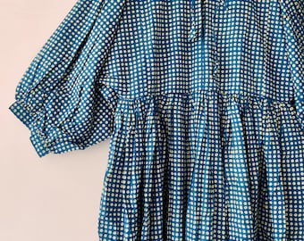 Puede incluir: Vestido azul con estampado de lunares blancos. El vestido tiene mangas largas abullonadas y cintura fruncida. Cuelga de una percha de madera sobre un fondo blanco.