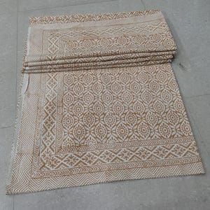 May include: Folded beige and white patterned fabric. The fabric features a repeating floral and geometric design. The border has a detailed pattern of triangles and floral motifs. The fabric is folded neatly, showcasing the intricate design.