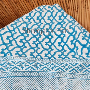 May include: White fabric with a blue and white geometric pattern. The fabric has a border with a repeating pattern of small diamonds and a chevron pattern at the bottom. The text "Shreejaipurfab" is printed in blue on the fabric.