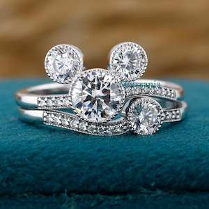 May include: A set of two silver rings with a Mickey Mouse-inspired design. The engagement ring features a large, round, clear gemstone, flanked by two smaller stones, and a band with smaller stones. The wedding band has a curved design with small stones.