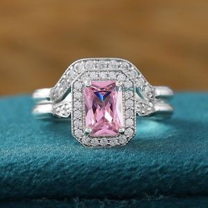 Könnte beinhalten: Ein silberner Ring mit einem rechteckigen rosafarbenen Edelstein, der von kleineren klaren Edelsteinen umgeben ist. Der Ring liegt auf einem blauen Samt-Hintergrund.