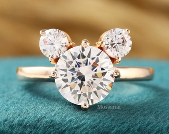 Puede incluir: Un anillo de oro con un gran diamante redondo en el centro y dos diamantes más pequeños a cada lado, formando una figura de Mickey Mouse.