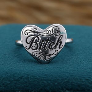 May include: A silver-toned ring featuring a heart-shaped face with the word Bitch engraved in a black script font. The heart is decorated with black filigree scrollwork patterns above and below the text. The brand name Moissania is visible.