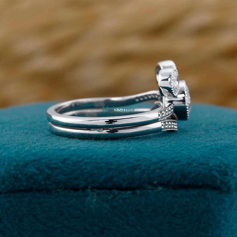 May include: A side view of a silver engagement ring set with a double band. The rings have a cluster of small, clear stones and a larger, round center stone. The rings are on a teal surface.