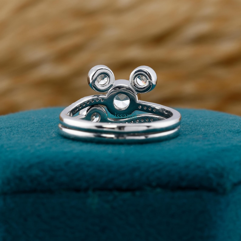 May include: A silver ring with a unique design featuring three prominent, round, clear gemstones. The ring has a double band and is set on a teal surface, with a blurred brown background.