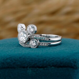 May include: A silver engagement ring set featuring a unique design. The main ring showcases a large, round, clear gemstone, with two smaller stones and two smaller round stones on the band. The design is reminiscent of a famous cartoon character.