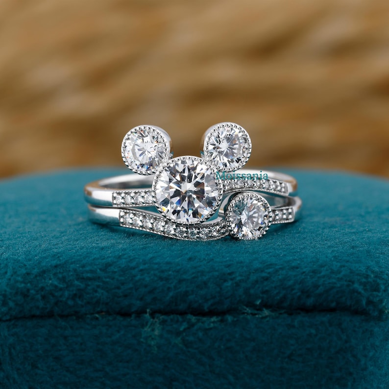 May include: A set of two silver rings with a Mickey Mouse-inspired design. The engagement ring features a large, round, clear gemstone, flanked by two smaller stones, and two smaller round stones on the band. The wedding band has a curved design with small gemstones.