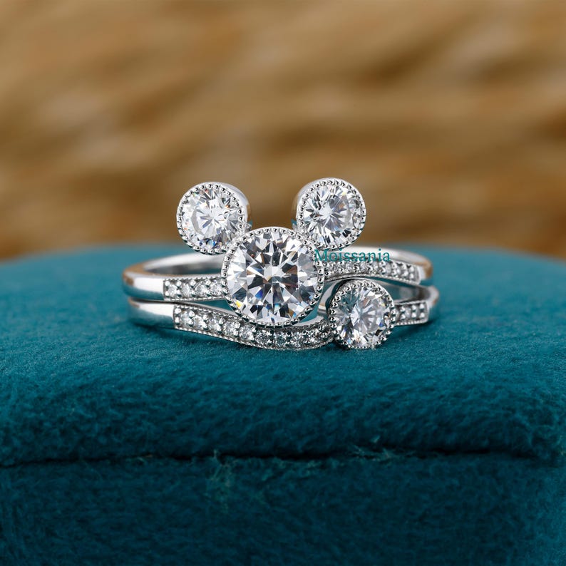May include: A set of two silver rings with a Mickey Mouse-inspired design. The engagement ring features a large, round, clear gemstone, flanked by two smaller stones, and a band with smaller stones. The wedding band has a curved design with small stones.