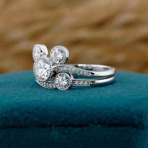 May include: A two-piece silver ring set featuring a Mickey Mouse-inspired design. The engagement ring has a large, round, clear gemstone with smaller stones around it. The band is adorned with smaller, clear gemstones. The wedding band is a simple, silver band.