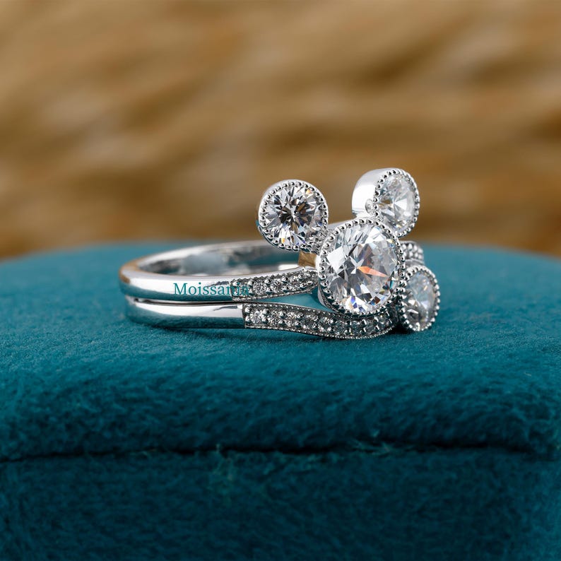 May include: A silver engagement ring set with a unique design. The ring features a large, round, clear gemstone in the center, with two smaller gemstones on either side, and two smaller gemstones above the center stone. The band is encrusted with smaller gemstones. The word "Moissanite" is engraved on the band.