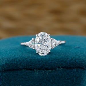 May include: A silver engagement ring with a large oval-cut center stone and two smaller triangular side stones. The ring is set on a blue velvet surface, creating a contrast that highlights the brilliance of the gemstones.
