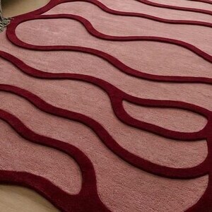 May include: A unique area rug with a flowing, organic design. The rug features a light pink central field with deep red, wave-like borders. The rug's shape is irregular, adding to its modern aesthetic. The rug is placed on a light wood floor.