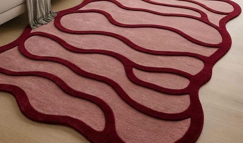 May include: A unique area rug with a flowing, organic design. The rug features a light pink central field with deep red, wave-like borders. The rug's shape is irregular, adding to its modern aesthetic. The rug is placed on a light wood floor.