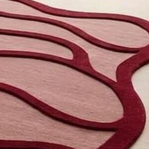 May include: A close-up of a rug with a unique, flowing design. The rug features a light pink central area with darker red outlines and accents. The edges of the rug are also a darker red, creating a layered effect.