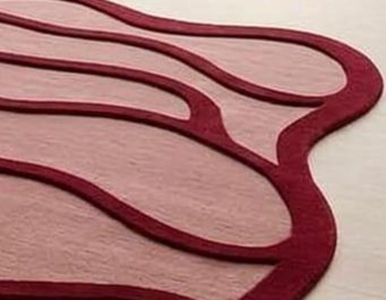 May include: A close-up of a rug with a unique, flowing design. The rug features a light pink central area with darker red outlines and accents. The edges of the rug are also a darker red, creating a layered effect.