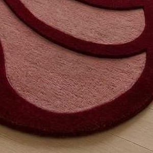 May include: Close-up of a burgundy and light red rug with a unique, abstract design. The rug has a plush texture and a contrasting border, creating a layered effect. The rug is placed on a light-colored wooden floor.