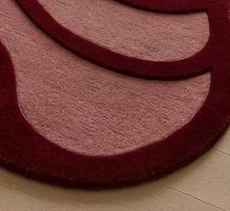 May include: Close-up of a burgundy and light red rug with a unique, abstract design. The rug has a plush texture and a contrasting border, creating a layered effect. The rug is placed on a light-colored wooden floor.