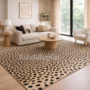 Puede incluir: Una alfombra con estampado de leopardo beige y negro en un salón. La alfombra es el punto focal, con un patrón repetido de manchas negras sobre un fondo beige. La habitación también cuenta con un sofá beige, una mesa de centro redonda de madera y una lámpara de araña.
