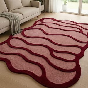 May include: A modern area rug with a unique, flowing design. The rug features a light pink central field with darker burgundy outlines and borders. The rug's shape is organic, with rounded edges and a soft texture, adding a touch of contemporary style to any living space.