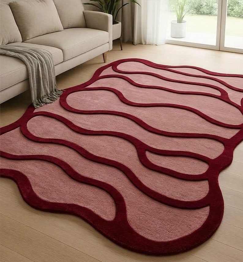 May include: A modern area rug with a unique, flowing design. The rug features a light pink central field with darker burgundy outlines and borders. The rug's shape is organic, with rounded edges and a soft texture, adding a touch of contemporary style to any living space.