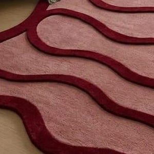 May include: A close-up of a rug with a wavy design. The rug features a light pink background with dark red, curved lines. The texture appears soft and plush, with a slight sheen. The design is modern and abstract.