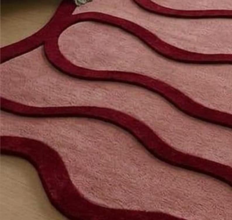 May include: A close-up of a rug with a wavy design. The rug features a light pink background with dark red, curved lines. The texture appears soft and plush, with a slight sheen. The design is modern and abstract.