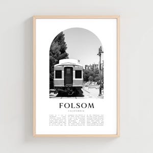 May include: Black and white framed print featuring a vintage train car under an archway. The print includes the text "FOLSOM CALIFORNIA" and a brief description of the city. The print is in a light wood frame.
